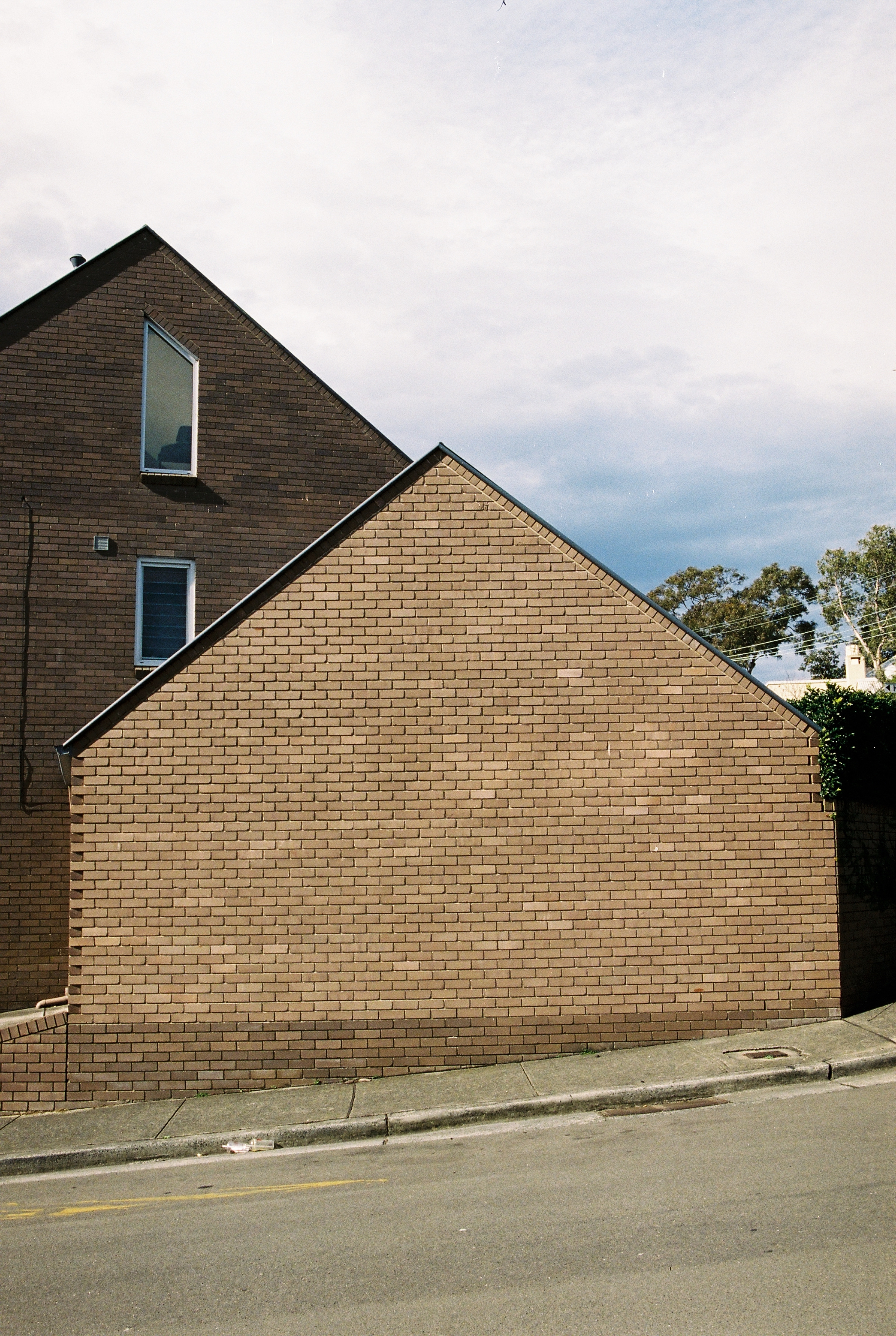 Exterior daytime view of a two-story brown brick house with an asymmetrical roof, windows, and a grey concrete sidewalk and street.