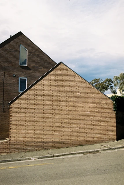 Exterior daytime view of a two-story brown brick house with an asymmetrical roof, windows, and a grey concrete sidewalk and street.