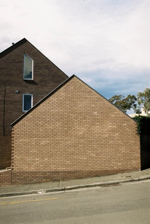 Exterior daytime view of a two-story brown brick house with an asymmetrical roof, windows, and a grey concrete sidewalk and street.