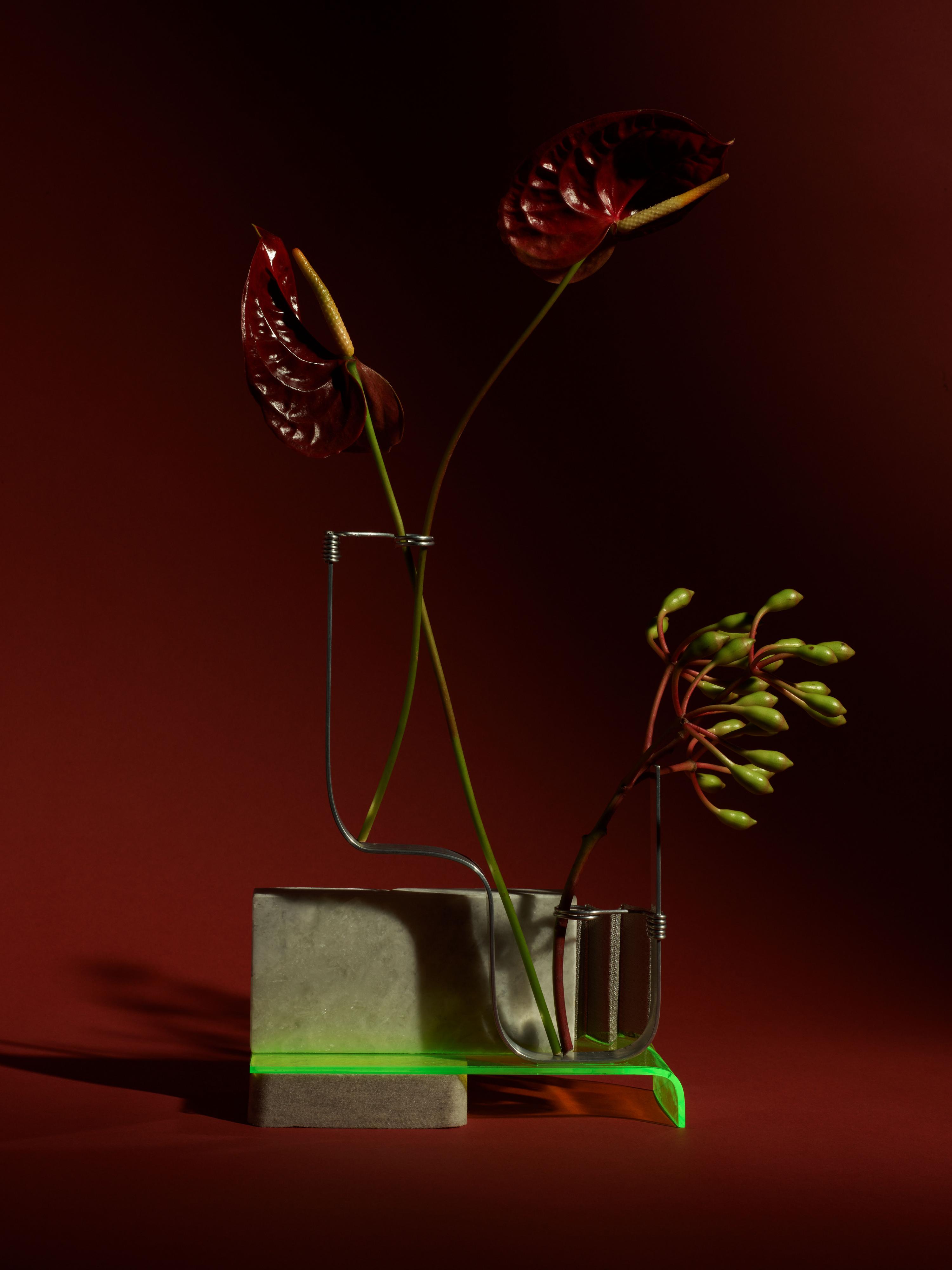 Still life of burgundy anthurium flowers in a modern vase set on a geometric green base against a solid burgundy background.