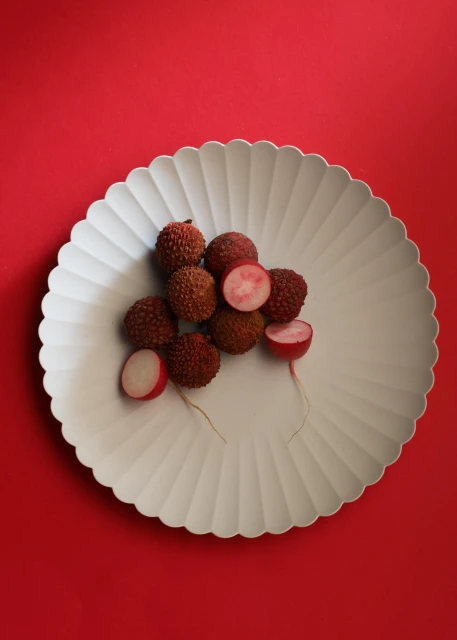 A pile of lychee fruits, some halved, arranged on a decorative white plate set against a vibrant red background in a studio setting.