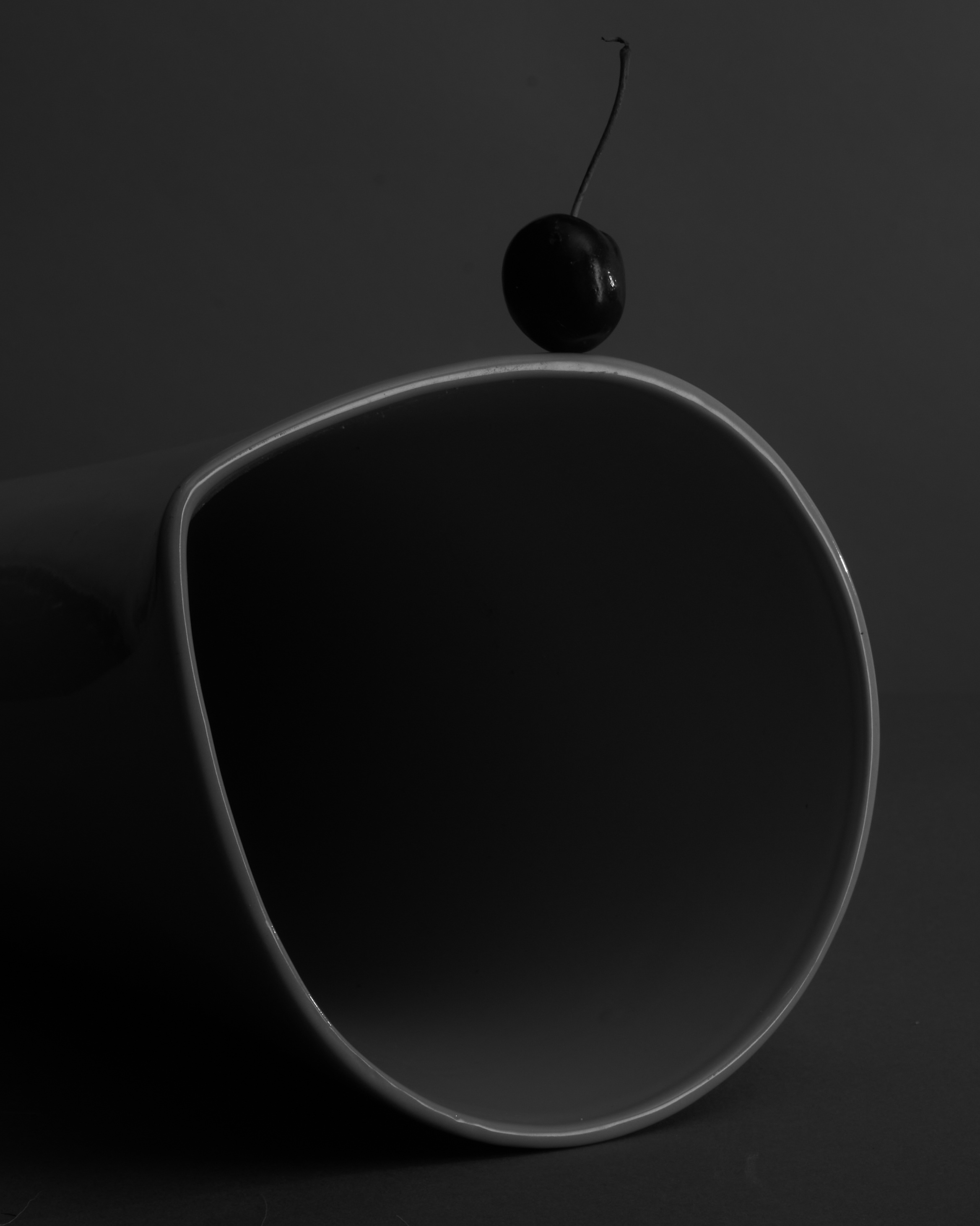 Monochromatic still life: a single cherry resting on the edge of a curved, glossy black surface, emphasizing simplicity and minimalism.