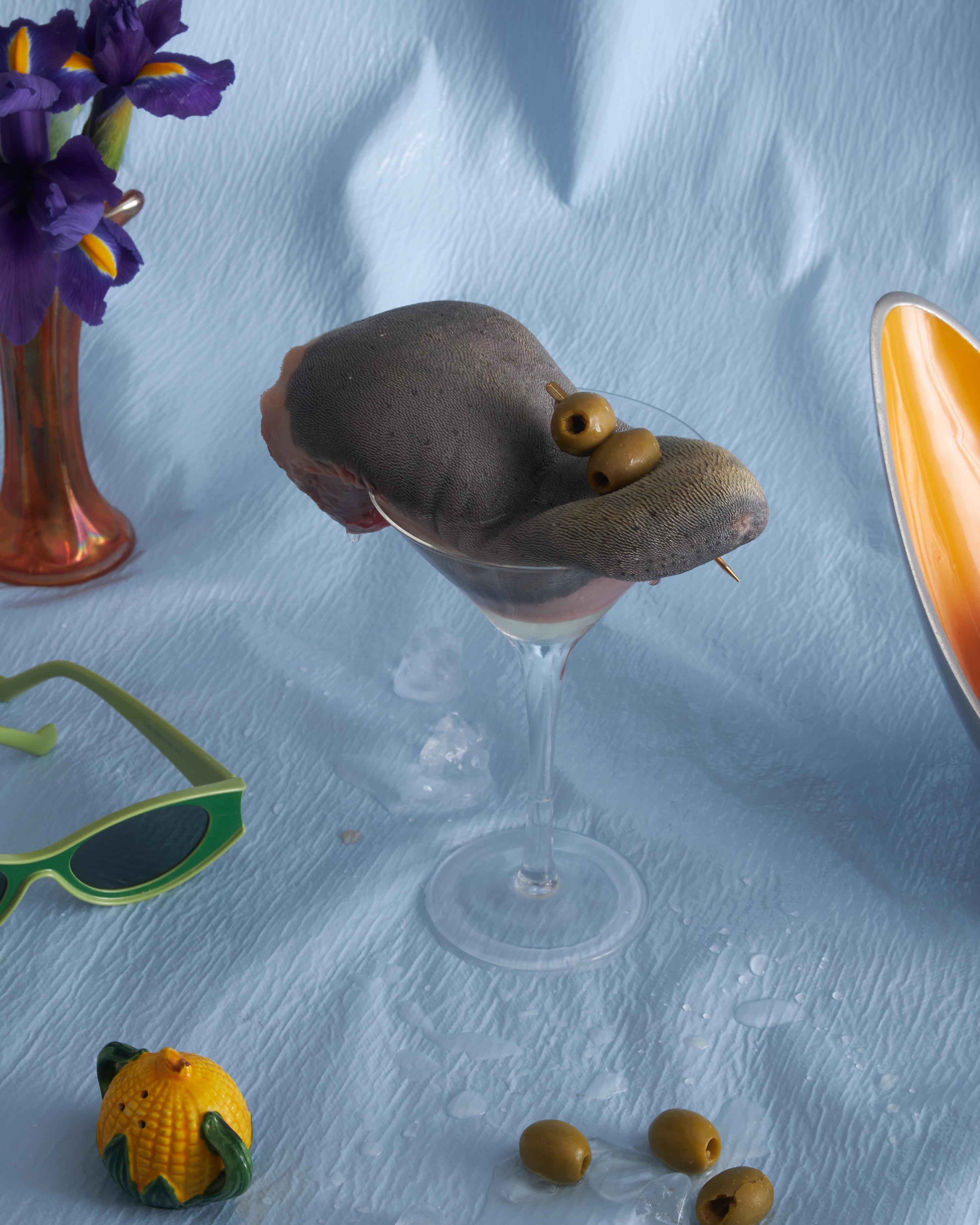 Still life with a cocktail glass topped with a hat and olives, set among sunglasses, a vase with irises, and a corn figurine.