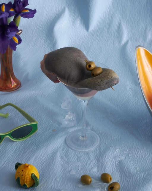 Still life with a cocktail glass topped with a hat and olives, set among sunglasses, a vase with irises, and a corn figurine.