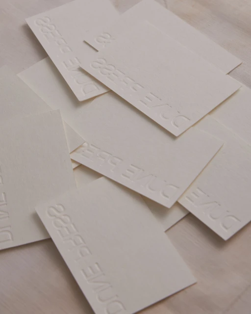 Stack of business cards with embossed text, arranged in a pile on a textured surface, creating a minimalist and tactile effect.