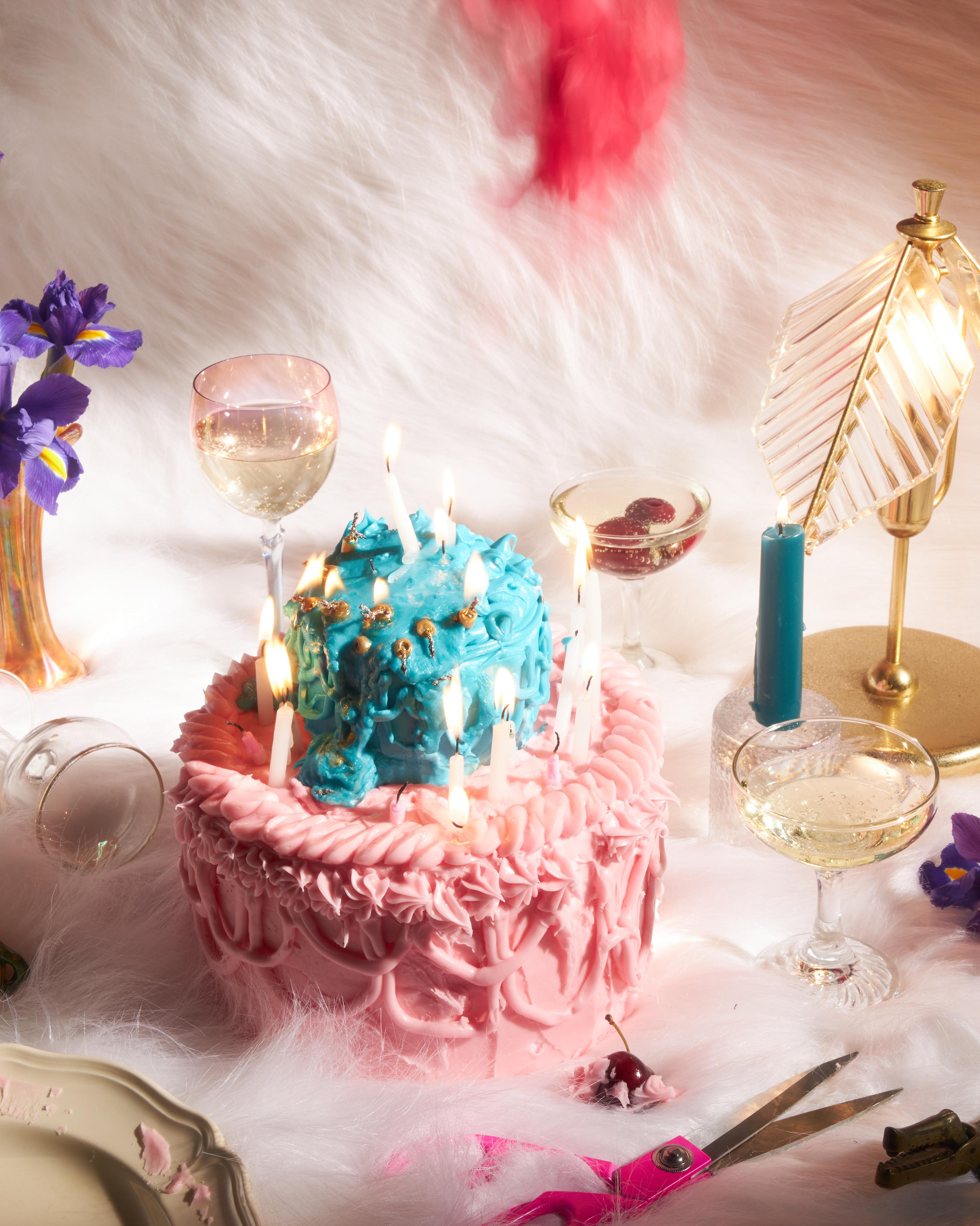 Celebration scene with a tiered birthday cake adorned with lit candles, surrounded by champagne glasses and floral decorations on a soft, white surface.