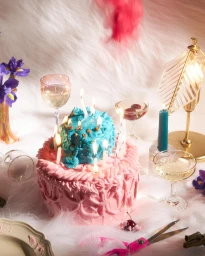 Birthday cake with pink frosting and a blue topper surrounded by lit candles, glassware, wine glasses, flowers, and a gold lamp