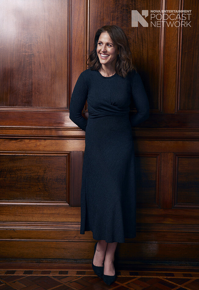 Portrait of a smiling woman in a long-sleeved dress standing indoors against a paneled wood wall, with neutral lighting.