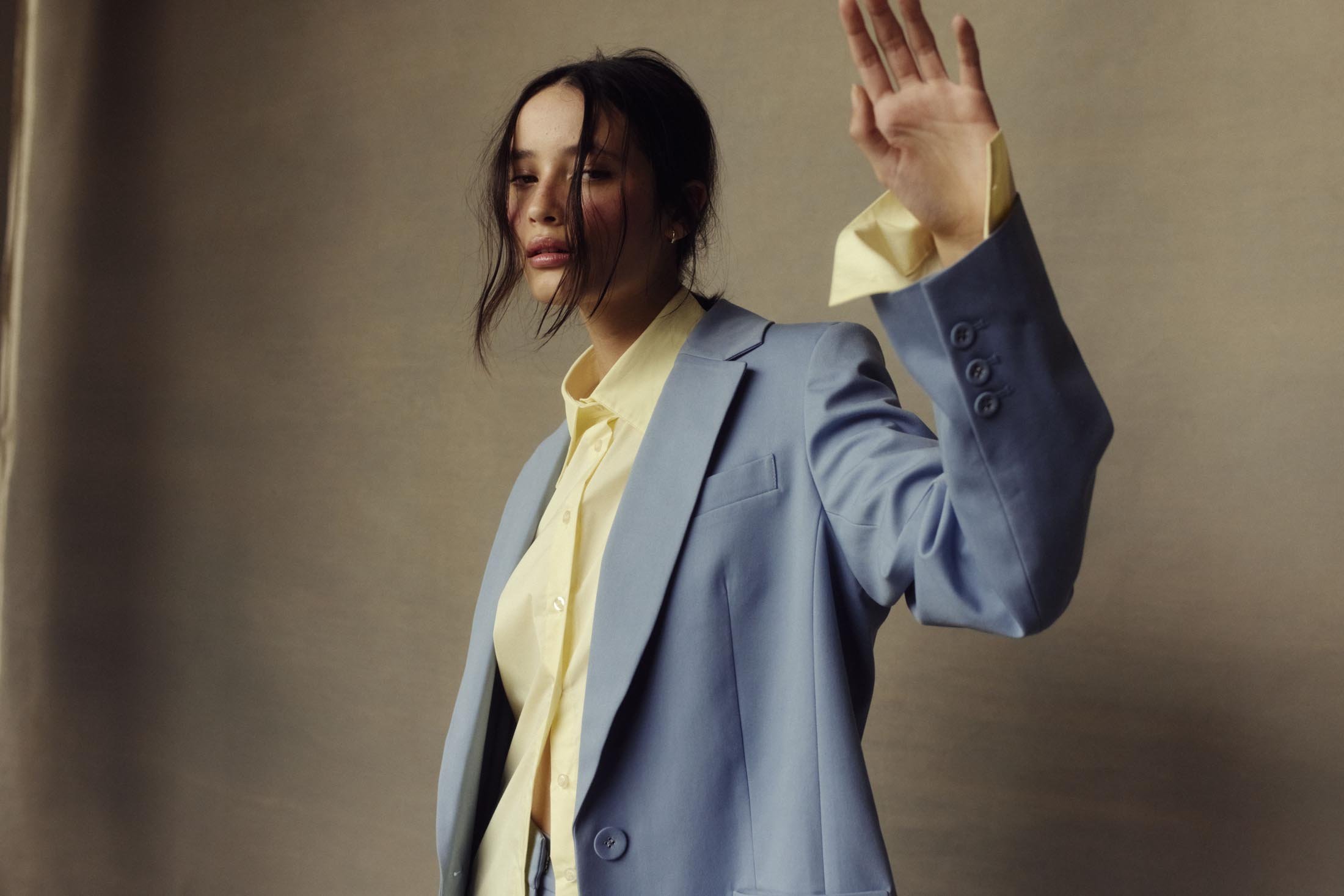 Woman in a light blue blazer and yellow collared shirt raises her hand in a fashion shot against a neutral gray backdrop.