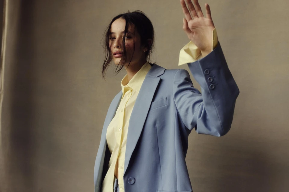 Woman in a light blue blazer and yellow collared shirt raises her hand in a fashion shot against a neutral gray backdrop.