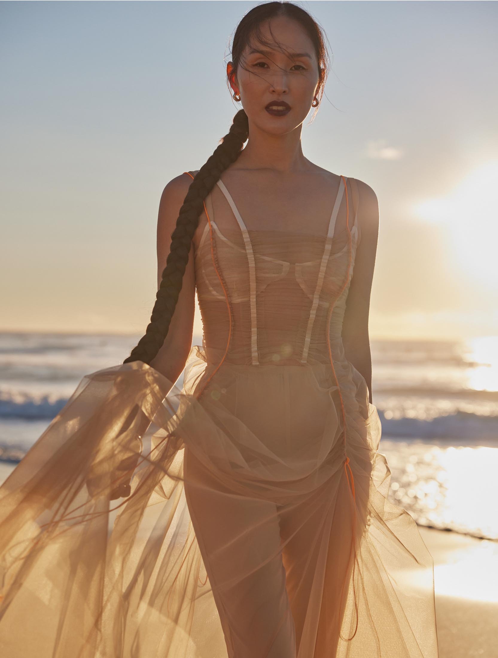 Woman in flowing dress poses on beach at sunset with long braid. Fashion photography with ocean and sky backdrop.