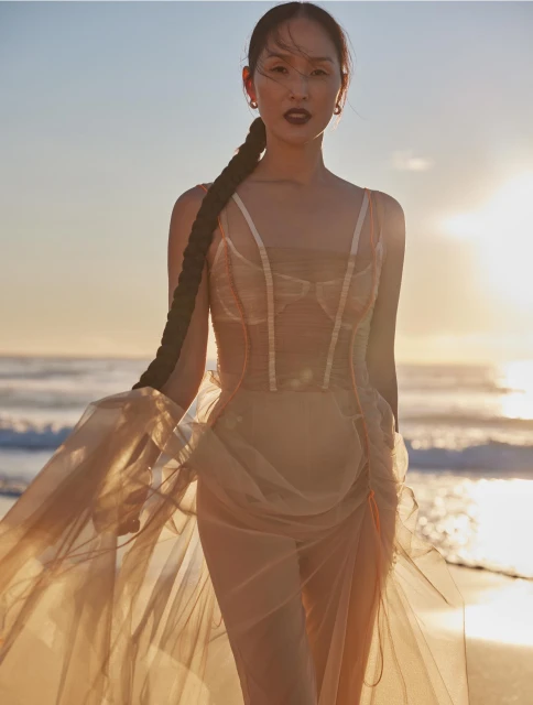 Woman in flowing dress poses on beach at sunset with long braid. Fashion photography with ocean and sky backdrop.