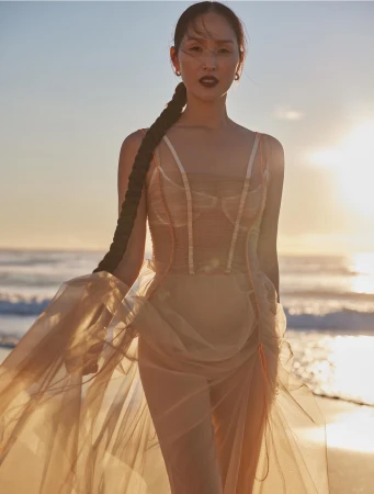 Woman in flowing dress poses on beach at sunset with long braid. Fashion photography with ocean and sky backdrop.