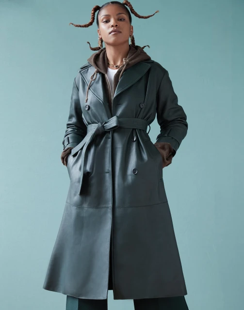 Portrait of a stylish adult woman wearing a belted green leather trench coat and brown hoodie against a light blue background.