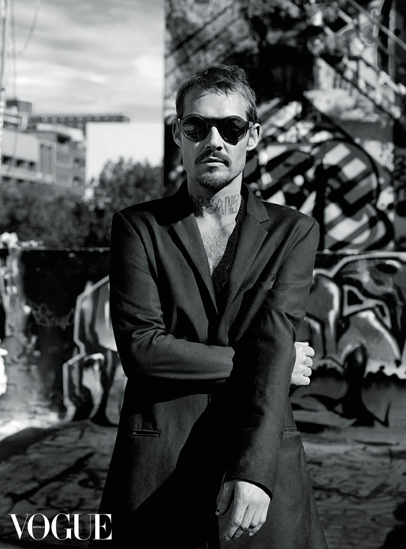Man wearing round sunglasses and a dark blazer standing against an urban graffiti background with Vogue watermark.