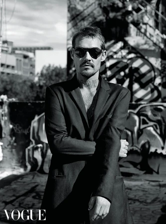 Man wearing round sunglasses and a dark blazer standing against an urban graffiti background with Vogue watermark.