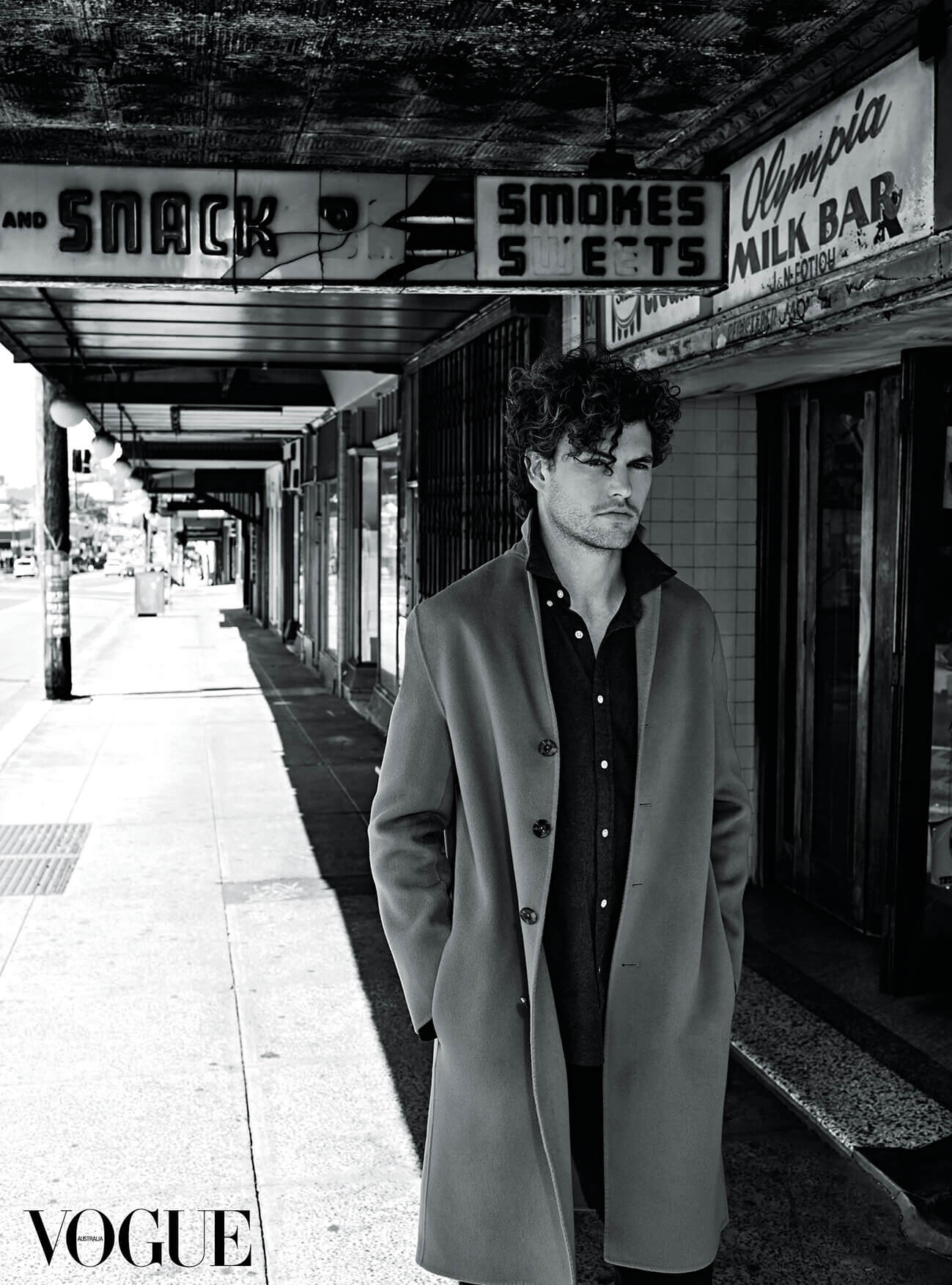 A man in a coat stands on a city sidewalk in a black and white photograph. The image is shot during daylight.