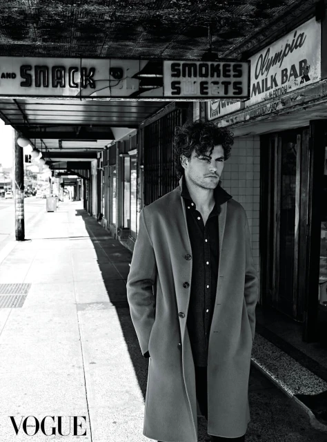 A man in a coat stands on a city sidewalk in a black and white photograph. The image is shot during daylight.