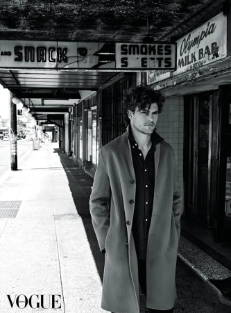 A man in a coat stands on a city sidewalk in a black and white photograph. The image is shot during daylight.