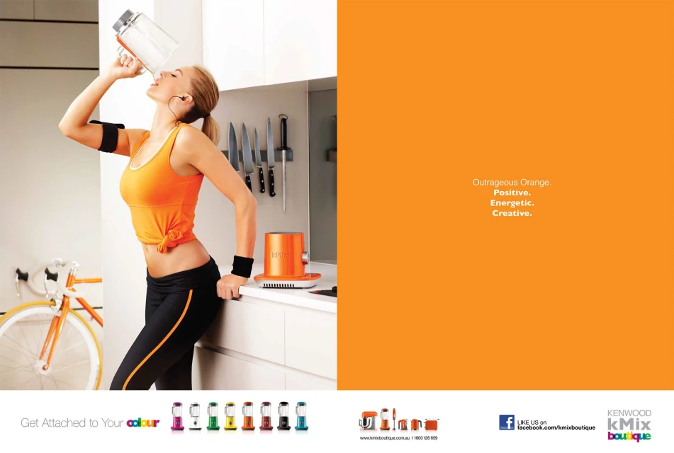 Blonde woman drinking from a large clear plastic bottle with an orange accent, wearing fitness clothing in a minimalist white kitchen.