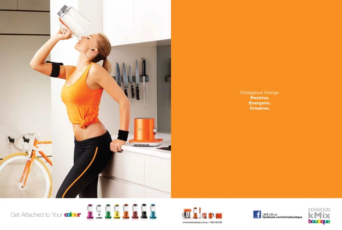 Blonde woman drinking from a large clear plastic bottle with an orange accent, wearing fitness clothing in a minimalist white kitchen.