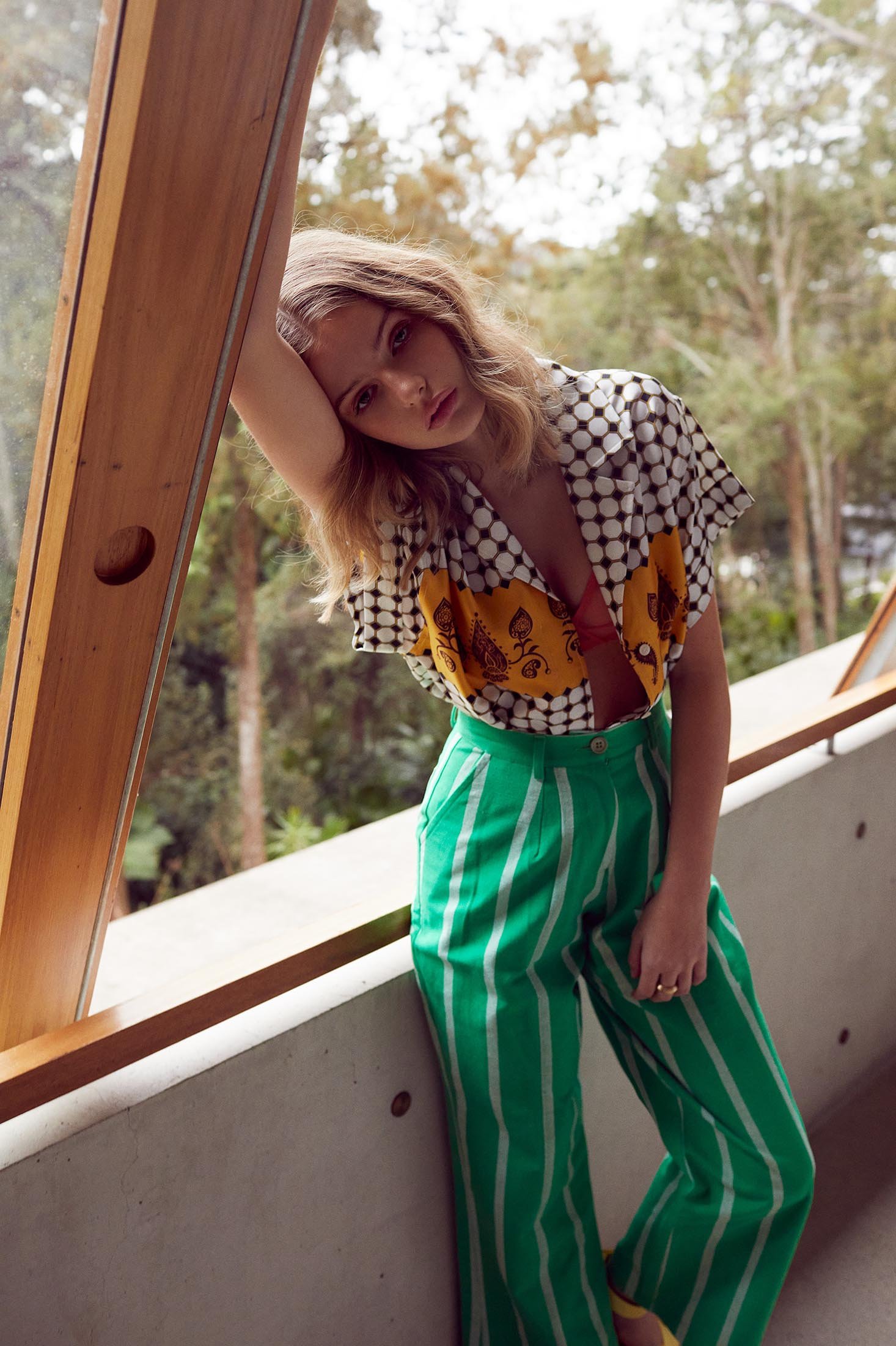A young woman in a patterned top and green striped pants leans against a window frame, with a blurred natural background.