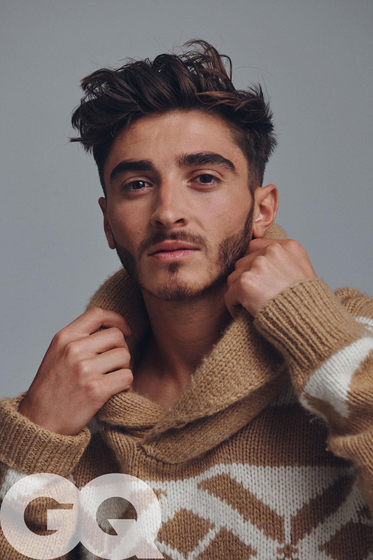 Portrait of a dark-haired male model wearing a brown and white patterned wool sweater with a neutral background and GQ logo.
