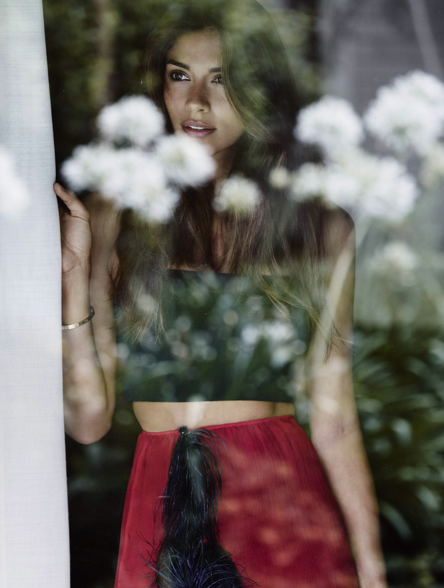 Woman with long brown hair poses in a red skirt and black top by a window with white flowers, looking to the left.