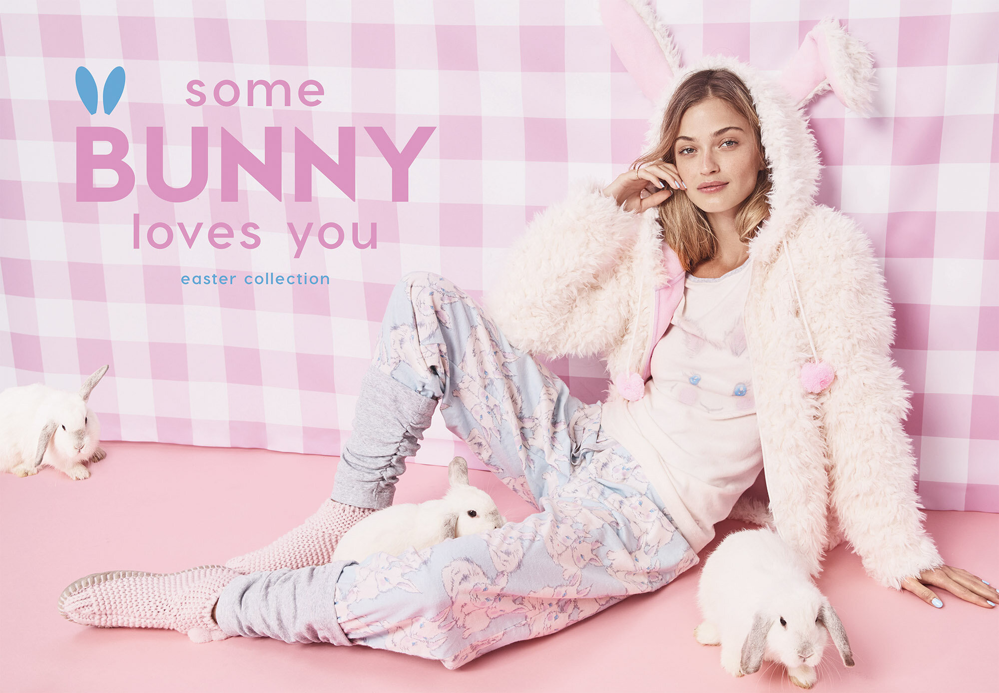 Woman wearing bunny ears and pajamas posing with white rabbits on pink backdrop in a studio setting for a fashion advertisement.