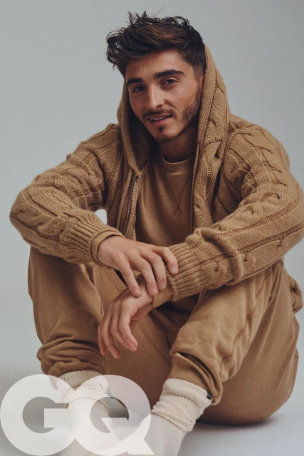 Portrait of a young adult male in a relaxed pose, wearing a tan knit hoodie and sweatpants, shot in a photo studio with soft, neutral lighting.