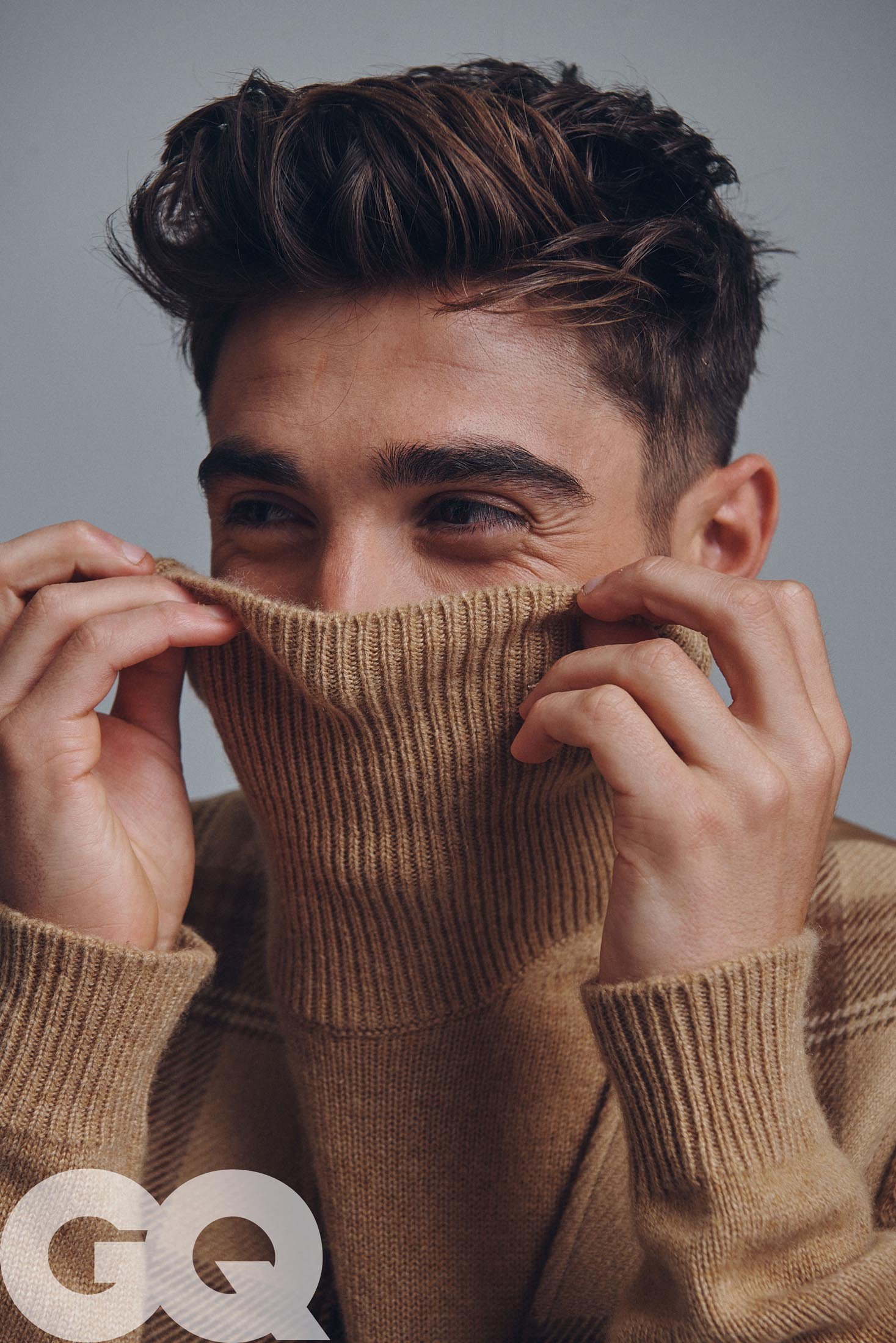 Portrait of a smiling adult male with short brown hair wearing a tan turtleneck sweater that covers part of his face. Gray background.