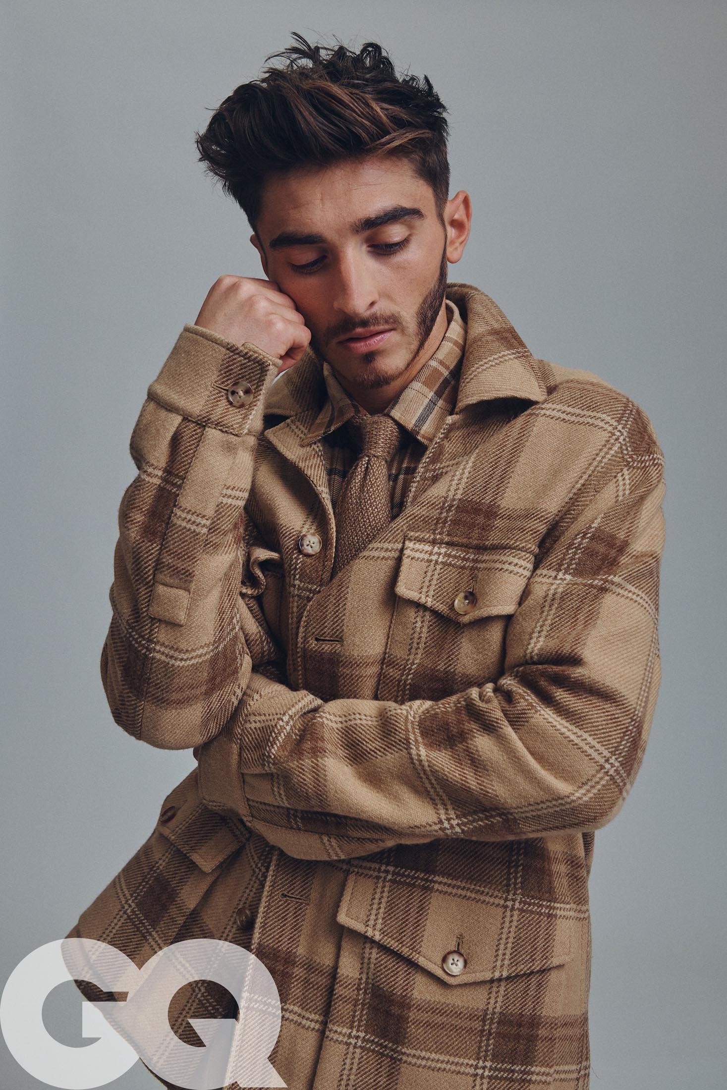 Portrait of a young adult male indoors wearing a patterned overshirt, looking downward with his hand touching his face.