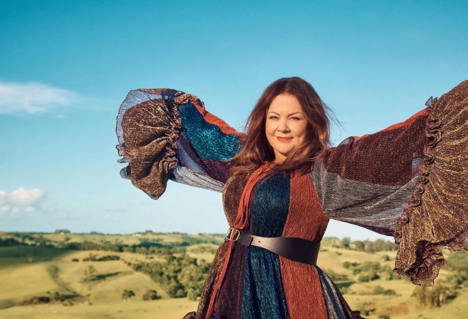 Woman with arms outstretched wearing a colorful sparkly long-sleeved dress against rolling green hills and a bright blue sky.