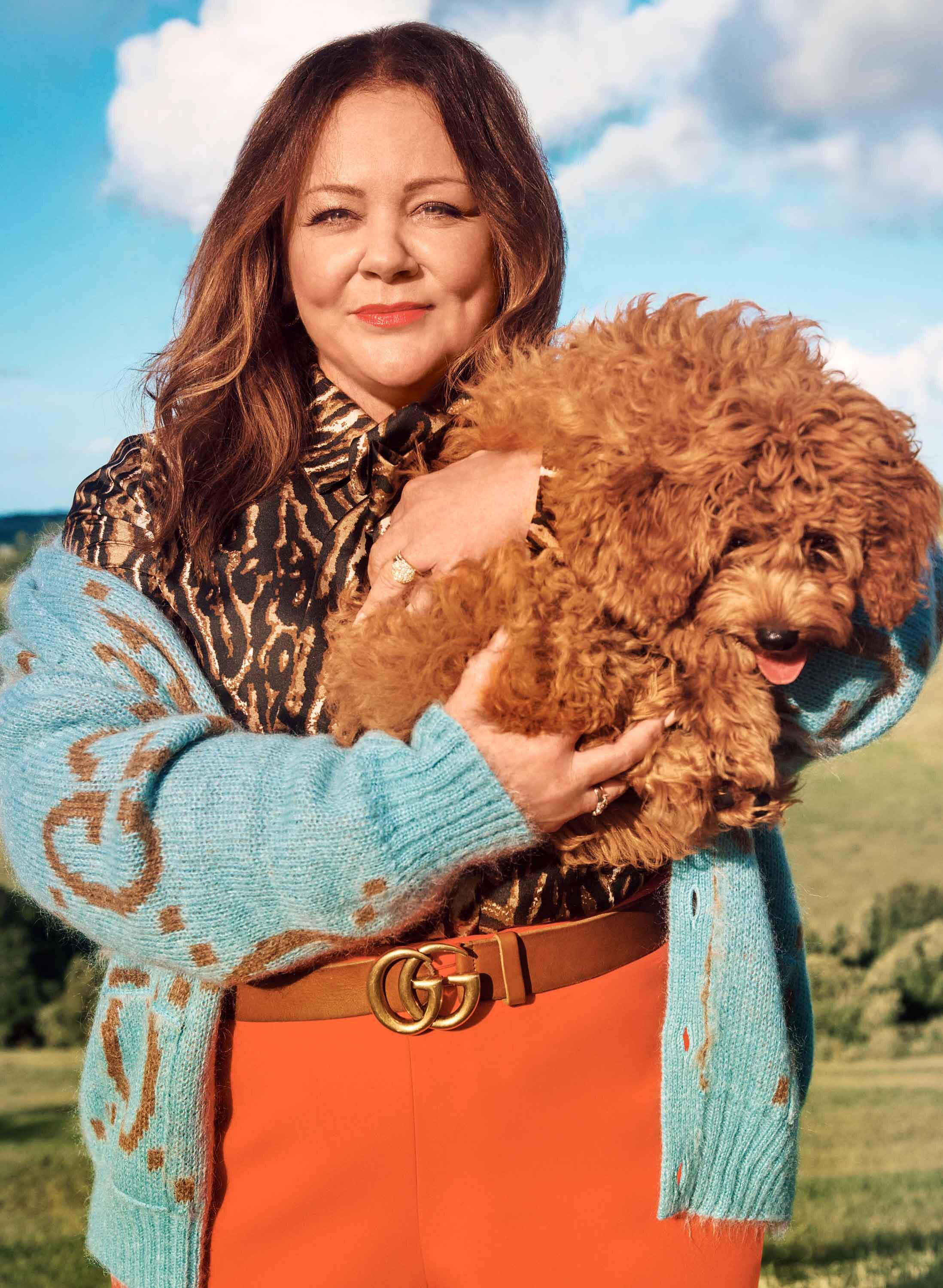 Melissa McCarthy poses outdoors with a brown poodle, wearing orange pants, a leopard-print blouse, and a blue cardigan with a Gucci belt.