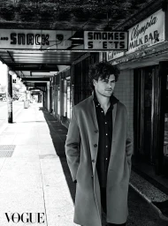 Monochrome portrait of a man with curly hair wearing a long coat standing on a city sidewalk beneath shop signs in Sydney.