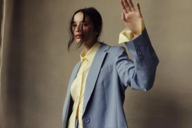 Model with dark hair wearing a light blue blazer over a yellow shirt raises a hand against a textured grey background.