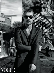 Man wearing round sunglasses and a dark blazer standing against an urban graffiti background with Vogue watermark.