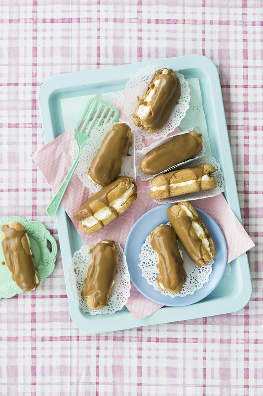 Overhead shot of cream-filled, caramel-iced éclairs arranged on a light blue tray with doilies and pink and mint green servingware.
