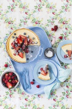 Overhead shot of a creamy tart topped with fresh berries, served on a blue platter with plates, forks, and floral tablecloth.