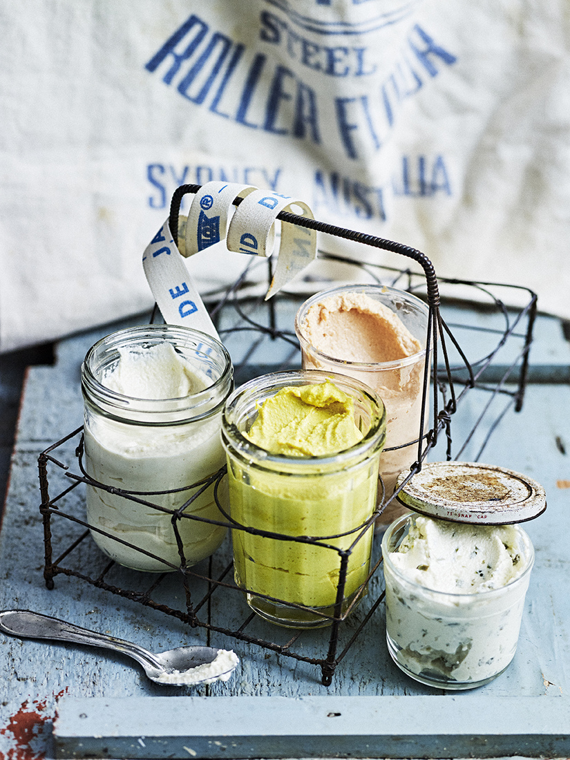 Four jars of different flavored dips are arranged in a wire basket, accompanied by a spoon on a rustic blue wooden surface.