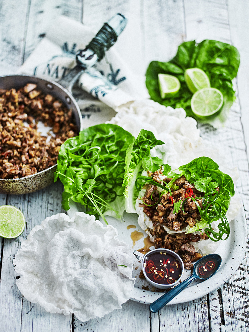 Overhead shot of lettuce wraps with seasoned meat, rice paper, sauce, and lime on a white wooden table, food photography.