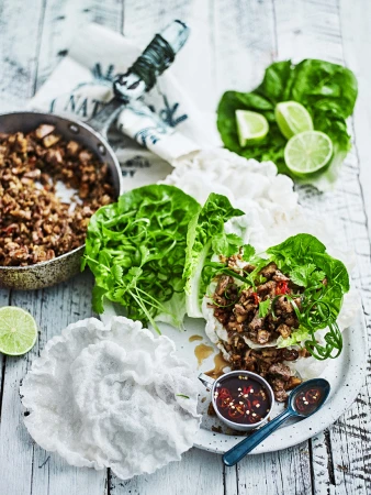 Overhead shot of lettuce wraps with seasoned meat, rice paper, sauce, and lime on a white wooden table, food photography.