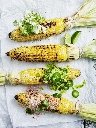 Overhead view of four ears of grilled corn on the cob, each topped with different ingredients, on white parchment paper.