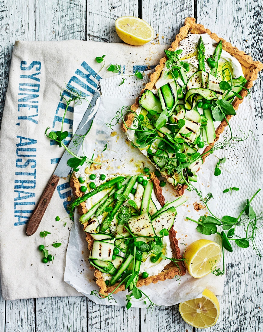 Overhead food styling shot of two savory tarts with asparagus and zucchini ribbons on a tea towel and white wooden tabletop.