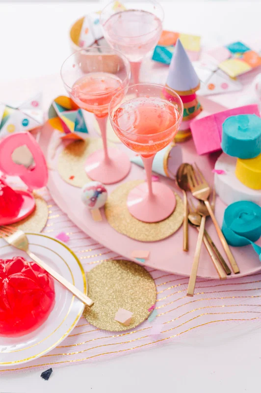 Overhead view of pink cocktails in stemmed glasses surrounded by party props and golden tableware on a pale pink surface.