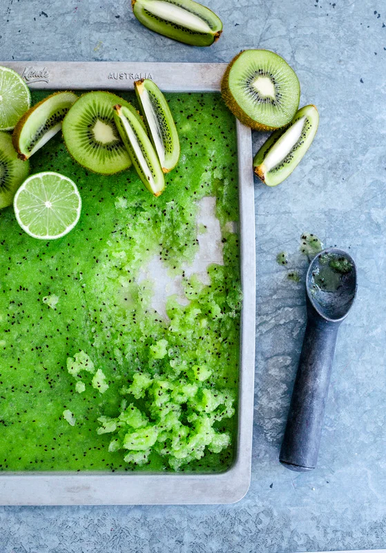Overhead food styling shot of vibrant green kiwi granita on a baking sheet with sliced kiwis, limes, and an ice cream scoop