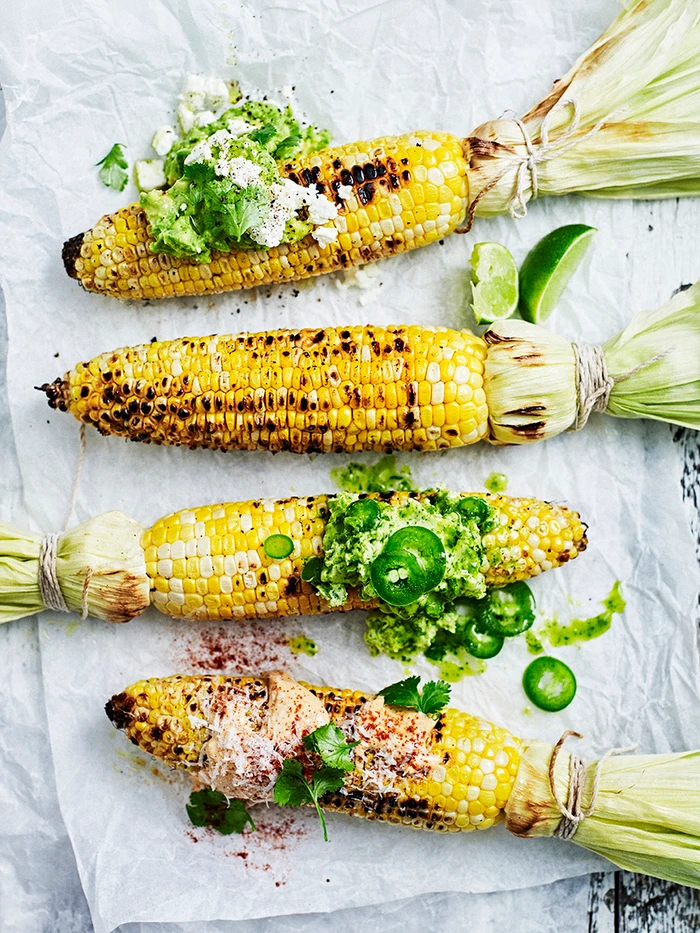 Four grilled corn cobs on crumpled parchment paper garnished with avocado, lime wedges, jalapeños, and fresh herbs.