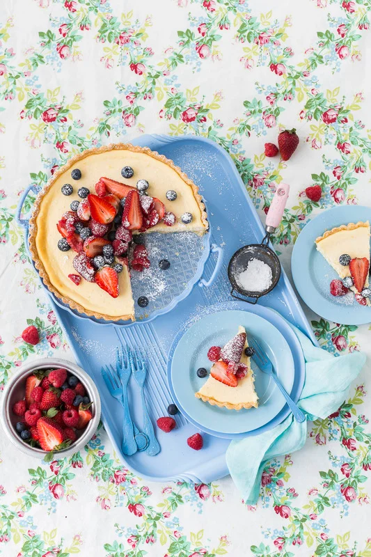 Overhead food styling shot of a berry tart on a blue tray with slices, fresh fruit, and a floral tablecloth.