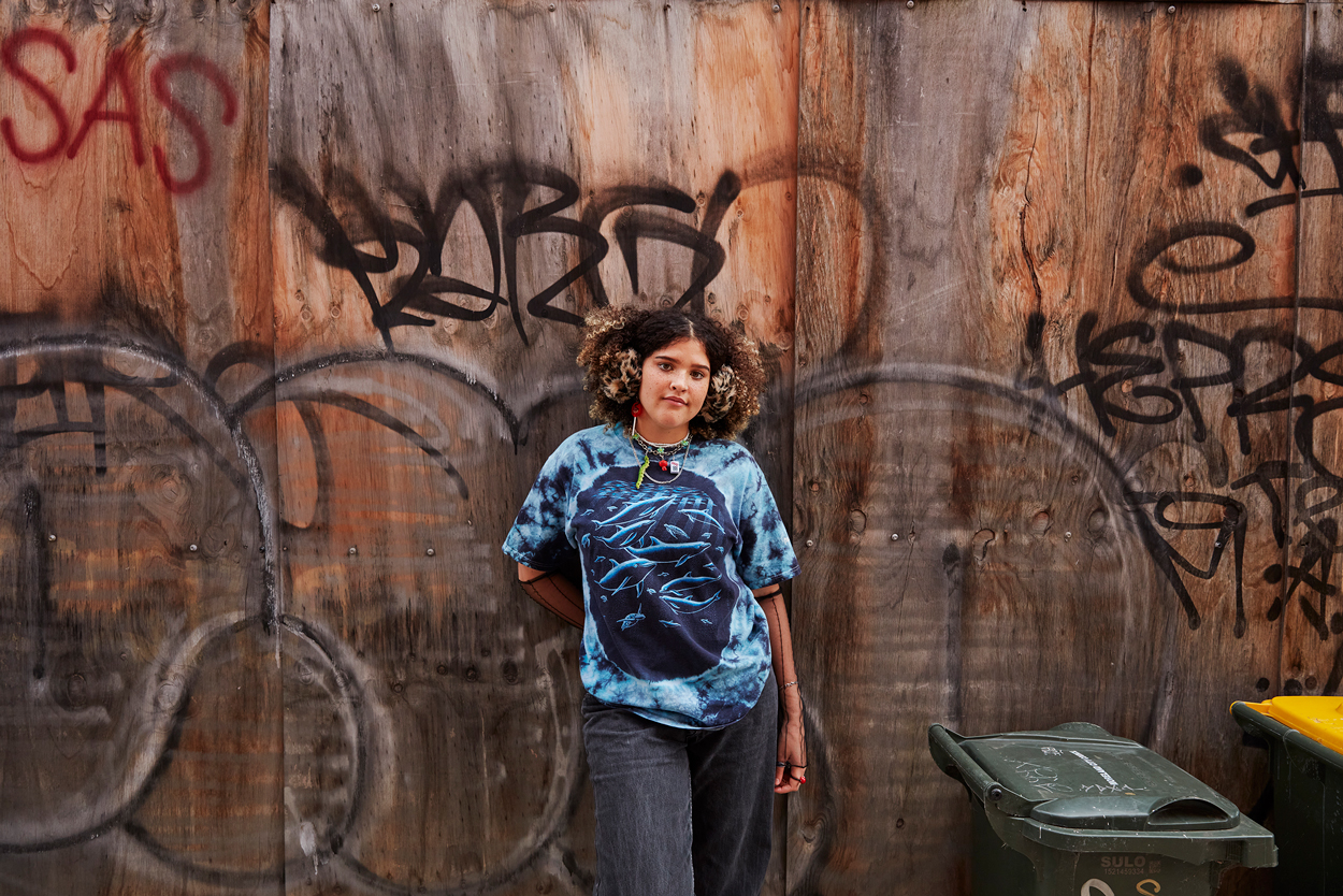 Young woman poses confidently in front of a graffiti-covered wooden wall, wearing a graphic tee and dark jeans in an outdoor setting.