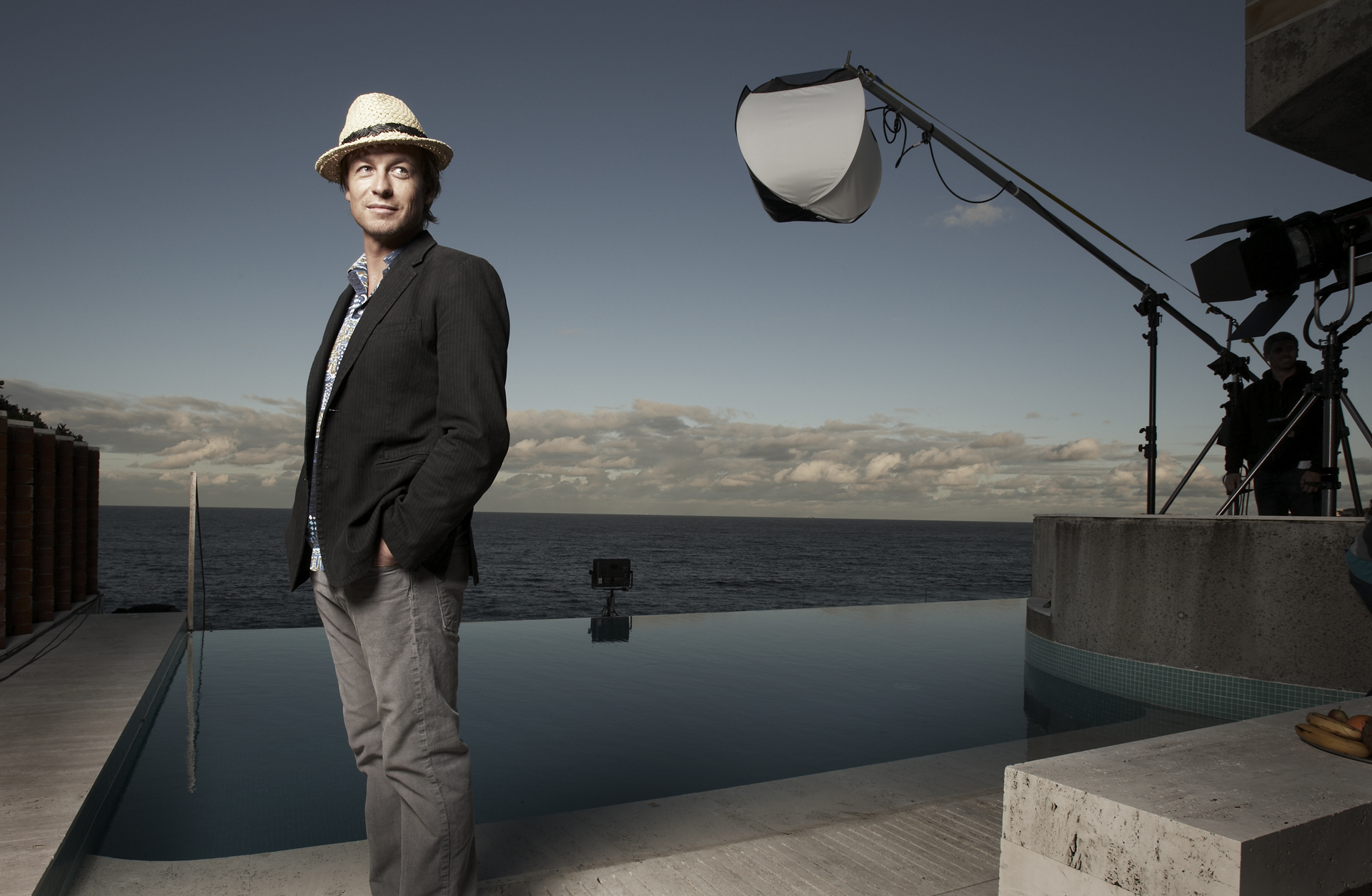 Adult male in a blazer and hat stands next to an infinity pool overlooking the ocean, with filming equipment and cloudy sky.